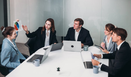 Business professionals engaged in meeting in conference room. Team members, dressed in formal attire, are discussing documents and using laptops, creating collaborative, productive work environment.の写真素材