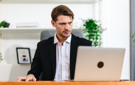 A skilled businessman sits at a desk, studying and analyzing financial reports while taking notes,...