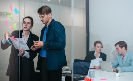 Businesspeople in a modern office, use sticky notes on glass wall for brainstorming and strategic planning. Idea organization, and project management in a corporate environment.の写真素材