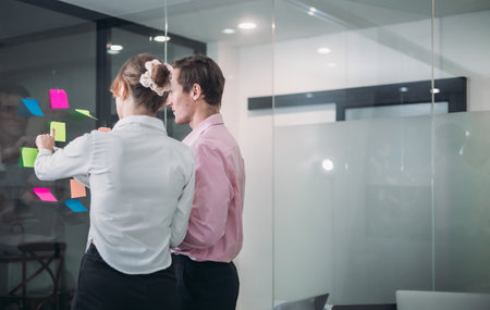 Businesspeople in a modern office, use sticky notes on glass wall for brainstorming and strategic planning. Idea organization, and project management in a corporate environment.の写真素材