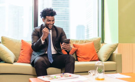 Businessperson sitting on couch in modern office space, engaged in video call on smartphone. The setting features warm decor with cushions and green plants, creating relaxed professional atmosphereの写真素材