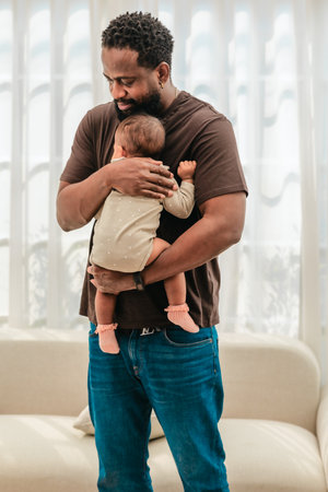A tender moment of a Black father gently holding his baby close, showcasing deep emotional connection and parental affection in a calm, softly lit home environment.の写真素材