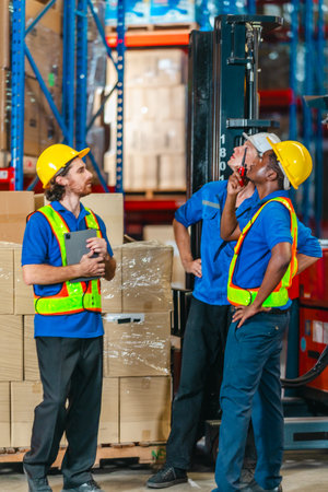 Three warehouse workers in safety vests and helmets coordinating logistics with a tablet and clipboard beside a forklift and pallet of boxes in an industrial storage facility.の写真素材