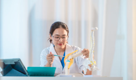 Asian medical student examines human anatomical models while referencing textbook and laptop, highlighting a detailed study session, medical education and hands-on learning in clinical setting.の写真素材