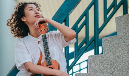 An Asian woman with curly, two-tone hair plays ukulele in an outdoor setting, radiating happiness and musical energy. A relaxed and artistic vibe with a natural and architectural elements backgroundの写真素材