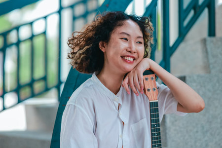 An Asian woman with curly, two-tone hair plays ukulele in an outdoor setting, radiating happiness and musical energy. A relaxed and artistic vibe with a natural and architectural elements backgroundの写真素材