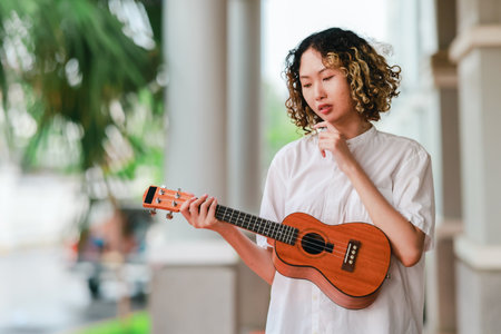 An Asian woman with curly, two-tone hair plays ukulele in an outdoor setting, radiating happiness and musical energy. A relaxed and artistic vibe with a natural and architectural elements backgroundの写真素材