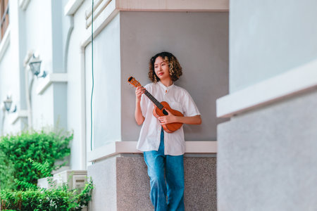 An Asian woman with curly, two-tone hair plays ukulele in an outdoor setting, radiating happiness and musical energy. A relaxed and artistic vibe with a natural and architectural elements backgroundの写真素材