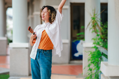 An Asian woman with curly, two-tone hair plays ukulele in an outdoor setting, radiating happiness and musical energy. A relaxed and artistic vibe with a natural and architectural elements backgroundの写真素材