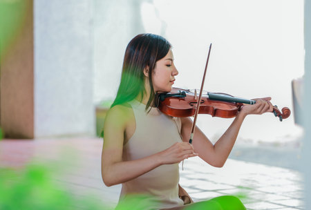 A young Asian woman gracefully practices the violin in a peaceful outdoor setting surrounded by lush greenery. Capturing a moment of musical concentration and tranquillity in nature.の写真素材