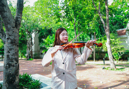 A young Asian woman gracefully practices the violin in a peaceful outdoor setting surrounded by lush greenery. Capturing a moment of musical concentration and tranquillity in nature.の写真素材
