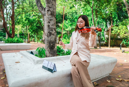 A young Asian woman gracefully practices the violin in a peaceful outdoor setting surrounded by lush greenery. Capturing a moment of musical concentration and tranquillity in nature.の写真素材