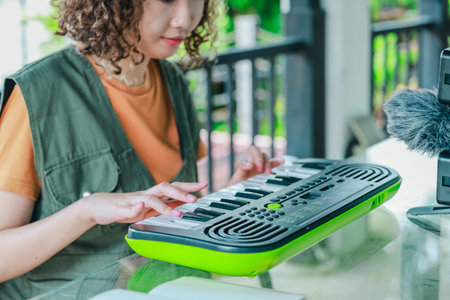 Asian woman with curly, two-tone hair engages in online music session using a small keyboard and smartphone, in a garden filled with blooming flowers. A moment of creative learning in outdoor settingの写真素材
