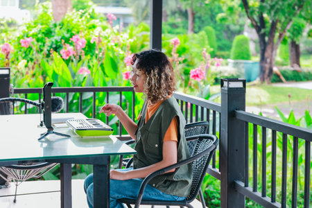 Asian woman with curly, two-tone hair engages in online music session using a small keyboard and smartphone, in a garden filled with blooming flowers. A moment of creative learning in outdoor settingの写真素材