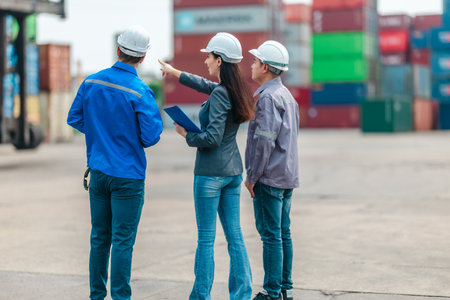 The female manager supervises and discusses logistics with two male workers at shipping container yard. Stacked containers and industrial equipment, highlight global trade and transportation industryの写真素材