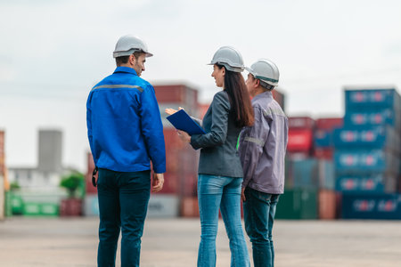 The female manager supervises and discusses logistics with two male workers at shipping container yard. Stacked containers and industrial equipment, highlight global trade and transportation industryの写真素材
