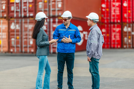 The female manager supervises and discusses logistics with two male workers at shipping container yard. Stacked containers and industrial equipment, highlight global trade and transportation industryの写真素材