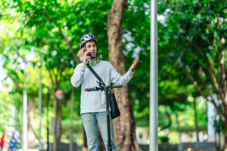 A young man wearing a helmet talks on his phone while standing with an electric scooter on a city street, capturing the essence of modern, eco-friendly urban transportation.の写真素材