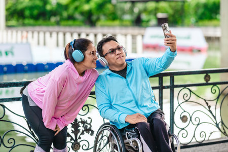 Cheerful couple in sporty casual outfits captures selfie moment together in outdoor park setting. One partner is seated in a wheelchair wearing headphones, while the other poses playfully beside himの写真素材