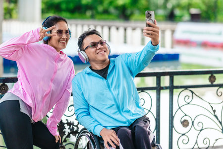 Cheerful couple in sporty casual outfits captures selfie moment together in outdoor park setting. One partner is seated in a wheelchair wearing headphones, while the other poses playfully beside himの写真素材