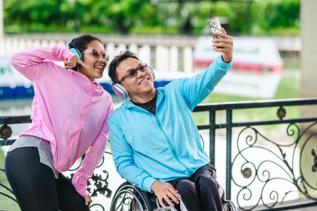 Cheerful couple in sporty casual outfits captures selfie moment together in outdoor park setting. One partner is seated in a wheelchair wearing headphones, while the other poses playfully beside himの写真素材