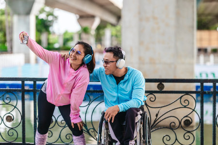 Cheerful couple in sporty casual outfits captures selfie moment together in outdoor park setting. One partner is seated in a wheelchair wearing headphones, while the other poses playfully beside himの写真素材