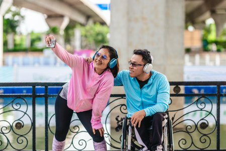 Cheerful couple in sporty casual outfits captures selfie moment together in outdoor park setting. One partner is seated in a wheelchair wearing headphones, while the other poses playfully beside himの写真素材