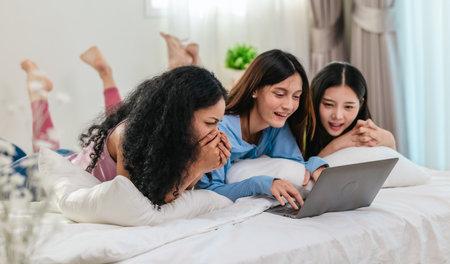 A group of diverse young women is lying on a bed, joyfully engaged in a digital game in a cosy, well-lit bedroom setting. The image captures friendship, leisure, fun and positive connection.の写真素材