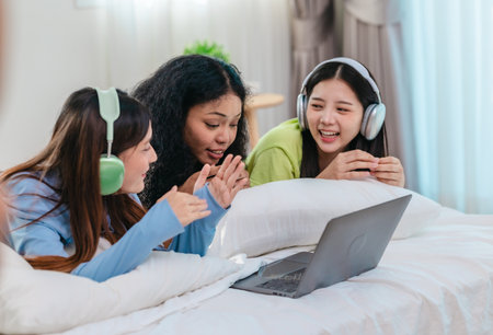 A group of diverse young women is lying on a bed, joyfully engaged in a digital game in a cosy, well-lit bedroom setting. The image captures friendship, leisure, fun and positive connection.の写真素材