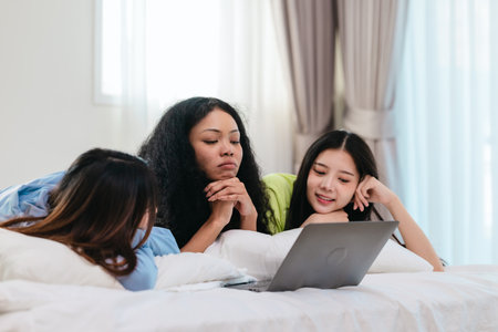 A group of diverse young women is lying on a bed, joyfully engaged in a digital game in a cosy, well-lit bedroom setting. The image captures friendship, leisure, fun and positive connection.の写真素材