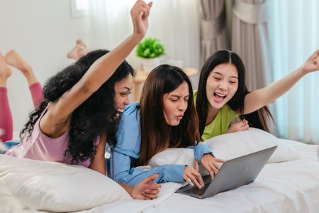 A group of diverse young women is lying on a bed, joyfully engaged in a digital game in a cosy, well-lit bedroom setting. The image captures friendship, leisure, fun and positive connection.の写真素材
