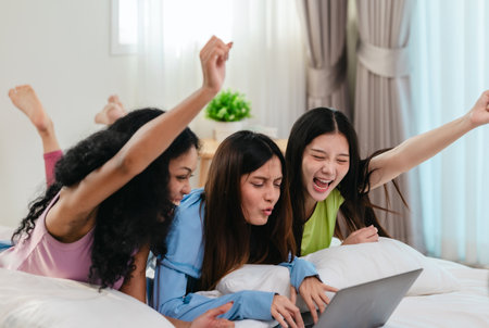 A group of diverse young women is lying on a bed, joyfully engaged in a digital game in a cosy, well-lit bedroom setting. The image captures friendship, leisure, fun and positive connection.の写真素材