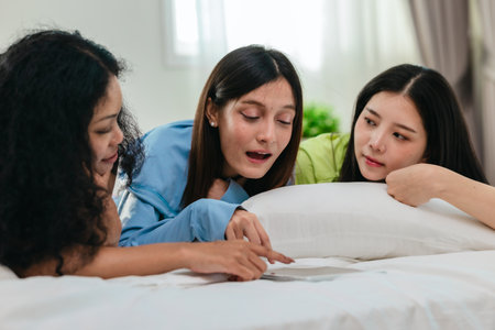 A group of diverse young women is lying on a bed, joyfully engaged in a digital game in a cosy, well-lit bedroom setting. The image captures friendship, leisure, fun and positive connection.の写真素材