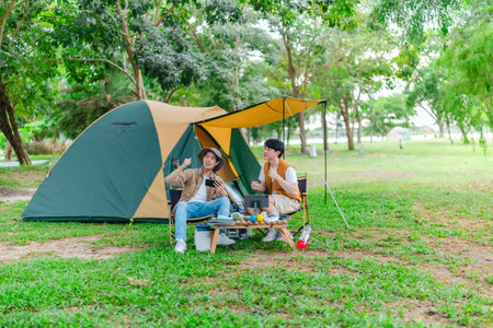 Two Asian male friends relax by the lake, enjoying music and singing together in serene outdoor setting. The scene captures joyful and peaceful moment of bonding and creativity during camping tripの写真素材