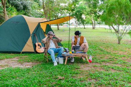 Two Asian male friends relax by the lake, enjoying music and singing together in serene outdoor setting. The scene captures joyful and peaceful moment of bonding and creativity during camping tripの写真素材