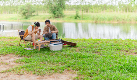 Two young Asian men enjoy playing video games together on tablet while relaxing at campsite. Surrounded by camping gear, guitars, and nature, the scene captures modern and joyful outdoor experience.の写真素材