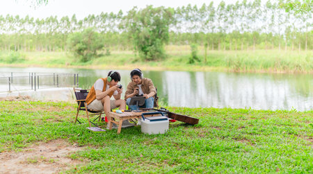 Two young Asian men enjoy playing video games together on tablet while relaxing at campsite. Surrounded by camping gear, guitars, and nature, the scene captures modern and joyful outdoor experience.の写真素材