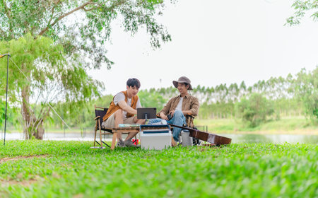 Two young Asian men enjoy playing video games together on tablet while relaxing at campsite. Surrounded by camping gear, guitars, and nature, the scene captures modern and joyful outdoor experience.の写真素材