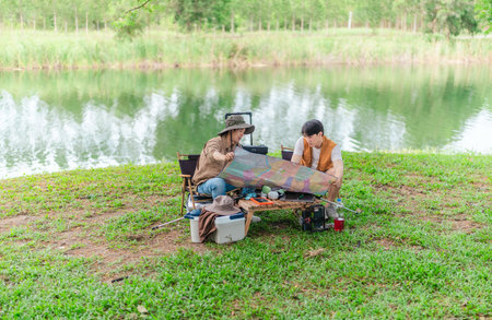 Two Asian male friends enjoy a calm camping experience beside a lake, surrounded by nature and gear....