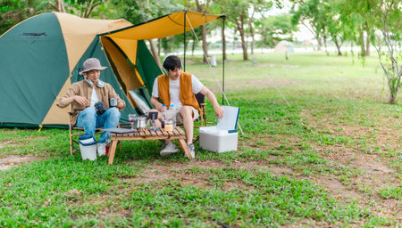 Two Asian male friends enjoy relaxing moment together at campsite surrounded by nature. With a tent set up and gear around them, the atmosphere reflects leisure, companionship, and outdoor freedomの写真素材