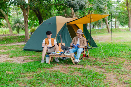 Two Asian male friends enjoy relaxing moment together at campsite surrounded by nature. With a tent set up and gear around them, the atmosphere reflects leisure, companionship, and outdoor freedomの写真素材