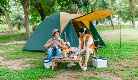 Two Asian male friends enjoy relaxing moment together at campsite surrounded by nature. With a tent...