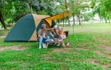 Two Asian male friends enjoy relaxing moment together at campsite surrounded by nature. With a tent set up and gear around them, the atmosphere reflects leisure, companionship, and outdoor freedomの写真素材