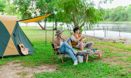 Two Asian male friends enjoy relaxing moment together at campsite surrounded by nature. With a tent set up and gear around them, the atmosphere reflects leisure, companionship, and outdoor freedomの写真素材