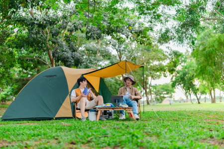 Two Asian male friends enjoy relaxing moment together at campsite surrounded by nature. With a tent set up and gear around them, the atmosphere reflects leisure, companionship, and outdoor freedomの写真素材