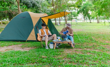 Two Asian male friends enjoy relaxing moment together at campsite surrounded by nature. With a tent set up and gear around them, the atmosphere reflects leisure, companionship, and outdoor freedomの写真素材