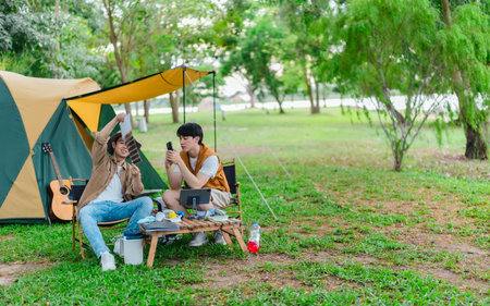 Two Asian male friends enjoy relaxing moment together at campsite surrounded by nature. With a tent set up and gear around them, the atmosphere reflects leisure, companionship, and outdoor freedomの写真素材