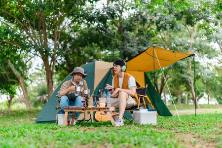 Two Asian male friends enjoy relaxing moment together at campsite surrounded by nature. With a tent set up and gear around them, the atmosphere reflects leisure, companionship, and outdoor freedomの写真素材
