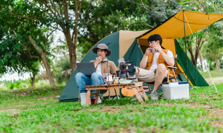 Two Asian male friends enjoy relaxing moment together at campsite surrounded by nature. With a tent set up and gear around them, the atmosphere reflects leisure, companionship, and outdoor freedomの写真素材