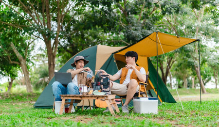 Two Asian male friends enjoy relaxing moment together at campsite surrounded by nature. With a tent set up and gear around them, the atmosphere reflects leisure, companionship, and outdoor freedomの写真素材